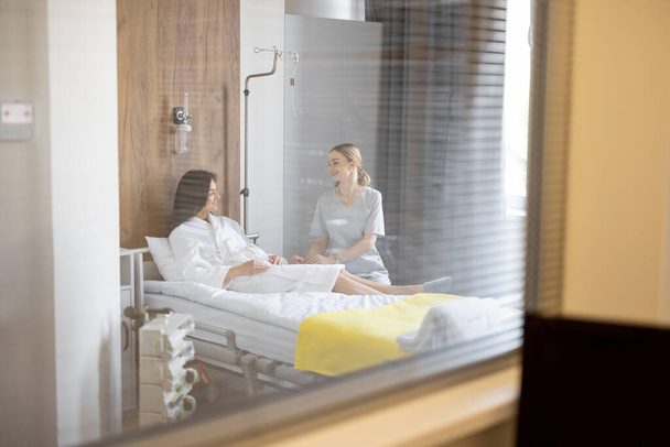 Nurse has a conversation with female patient lying in bed during rehabilitation or medical treatment in a ward. View from a surveillance room - Фото, зображення