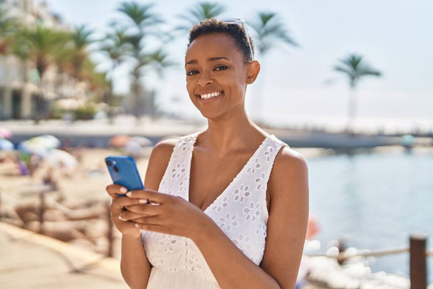 Femme afro-américaine souriante confiante en utilisant un smartphone au bord de la mer - Photo, image