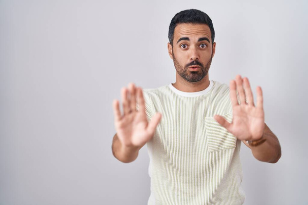 Hispanic man with beard standing over isolated background moving away hands palms showing refusal and denial with afraid and disgusting expression. stop and forbidden.  - Foto, imagen