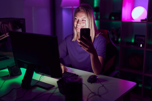 Молодая блондинка стример с помощью компьютера и смартфона в игровой комнате - Фото, изображение