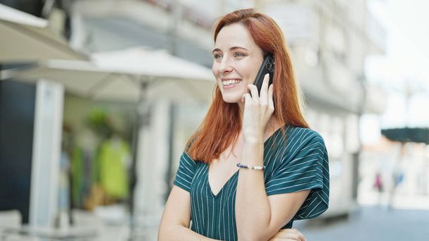 Genç kızıl saçlı, gülümseyen, kendine güvenen bir kadın kahve dükkanının terasında akıllı telefondan konuşuyor. - Fotoğraf, Görsel