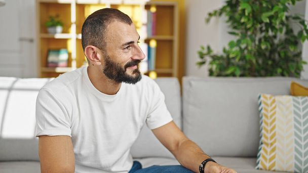 Junger hispanischer Mann lächelt selbstbewusst auf Sofa sitzend und spricht zu Hause - Foto, Bild