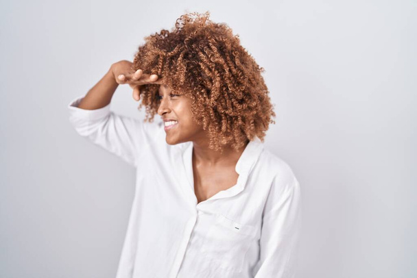 Young hispanic woman with curly hair standing over white background very happy and smiling looking far away with hand over head. searching concept.  - Foto, afbeelding