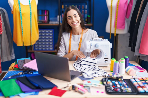Молода красива іспаномовна жінка кравець використовує швейну машину і ноутбук на фабриці одягу
 - Фото, зображення
