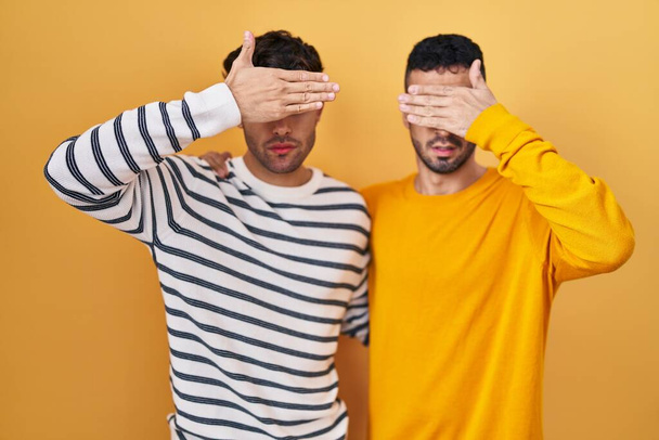 Giovane coppia gay ispanica in piedi su sfondo giallo che copre gli occhi con la mano, guardando serio e triste. concetto di avvistamento, nascondiglio e rifiuto  - Foto, immagini