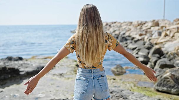 Young blonde woman standing with arms open backwards at seaside - Fotoğraf, Görsel