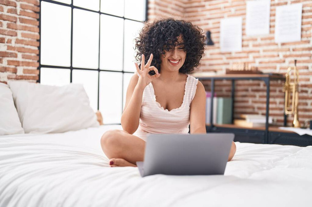 Молодая женщина с Ближнего Востока с помощью компьютера ноутбука на кровати делает хорошо знак с пальцами, улыбаясь дружелюбный жест отличный символ  - Фото, изображение