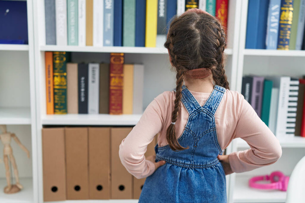 Adorable hispanic girl student looking for book standing backwards at classroom - Foto, Imagem