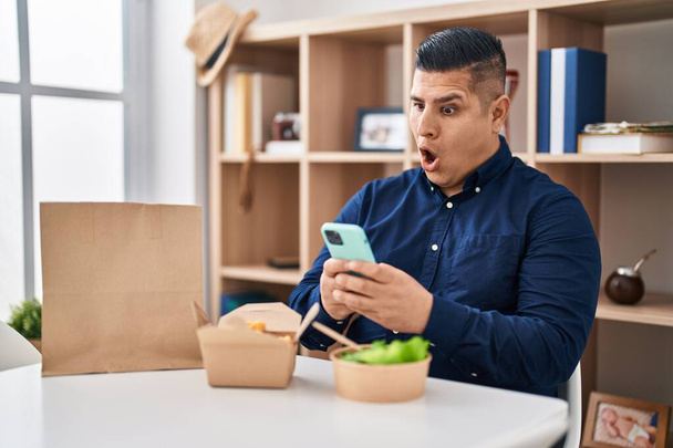 İspanyol genç adam yemek yiyor. Şok suratlı akıllı telefon kullanıyor. Şüpheci ve alaycı görünüyor.  - Fotoğraf, Görsel