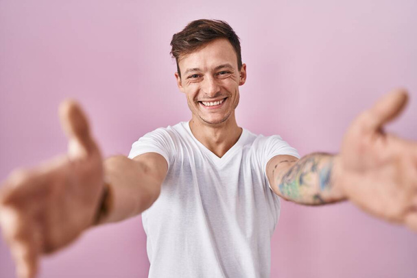 Fehér férfi áll a rózsaszín háttér felett, és a kamerát nézi, ahogy tárt karokkal mosolyog. vidám kifejezés, mely felöleli a boldogságot.  - Fotó, kép