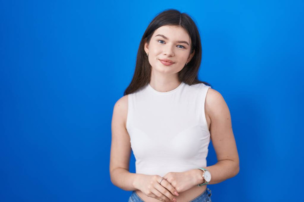 Young caucasian woman standing over blue background with hands together and crossed fingers smiling relaxed and cheerful. success and optimistic  - 写真・画像