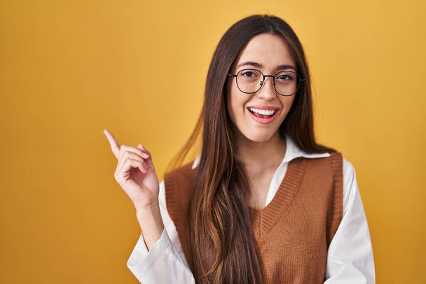 Young brunette woman standing over yellow background wearing glasses with a big smile on face, pointing with hand and finger to the side looking at the camera.  - Φωτογραφία, εικόνα