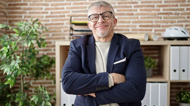 Μεσήλικας γκριζομάλλης επιχειρηματίας χαμογελά αυτοπεποίθηση στέκεται με τα χέρια σταυρωμένα χειρονομία στο γραφείο - Φωτογραφία, εικόνα