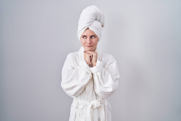 Blonde caucasian woman wearing bathrobe laughing nervous and excited with hands on chin looking to the side  - Foto, Bild