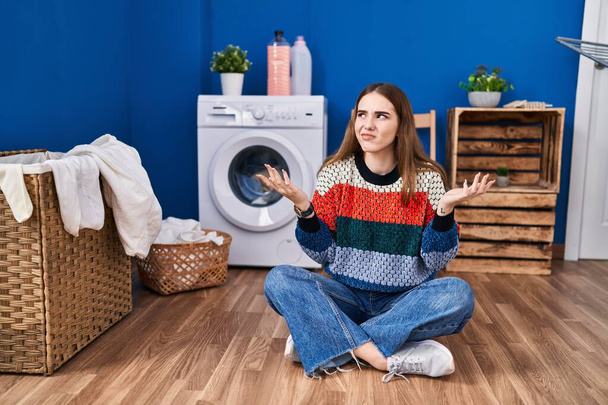 Young hispanic girl doing laundry clueless and confused expression with arms and hands raised. doubt concept.  - Photo, Image