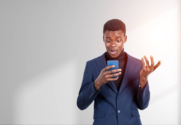 Handsome African American businessman wearing formal suit is standing watching at smartphone near empty white wall in background. Concept of modern gadgets, mobile communication, time management - Photo, image