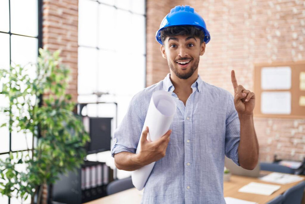 Homme arabe avec barbe portant un casque d'architecte au bureau de construction surpris par une idée ou une question pointant du doigt avec un visage heureux, numéro un  - Photo, image