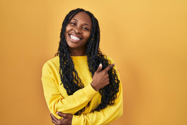 Donna africana in piedi su sfondo giallo con un grande sorriso sul viso, puntando con mano e dito verso il lato guardando la fotocamera.  - Foto, immagini
