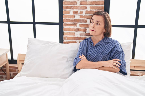 Mulher de meia idade sentada na cama com expressão infeliz e braços cruzados gesto no quarto - Foto, Imagem