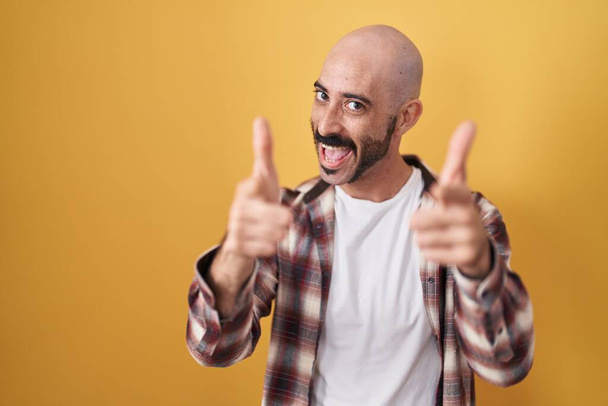 幸福と面白い顔を持つカメラに指を指して黄色の背景に立ってひげを持つヒスパニック系の男。良いエネルギーと振動は.  - 写真・画像