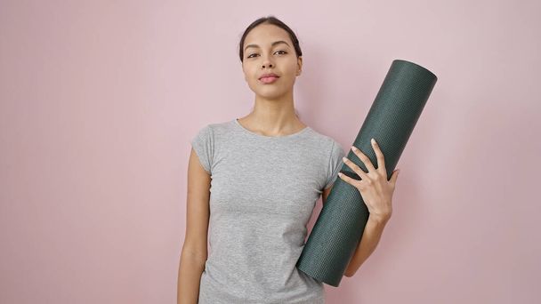 Jonge mooie latino vrouw dragen sportkleding houden yoga mat over geïsoleerde roze achtergrond - Foto, afbeelding