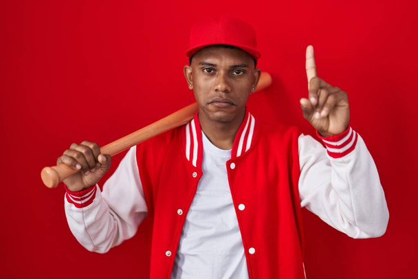 Young hispanic man playing baseball holding bat pointing up looking sad and upset, indicating direction with fingers, unhappy and depressed.  - Foto, immagini
