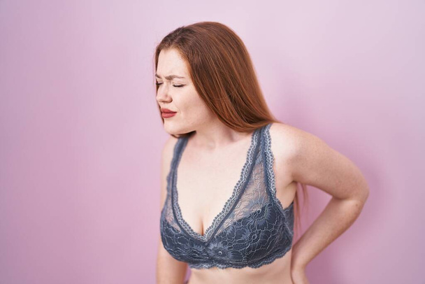 Redhead woman wearing lingerie over pink background suffering of backache, touching back with hand, muscular pain  - Photo, Image
