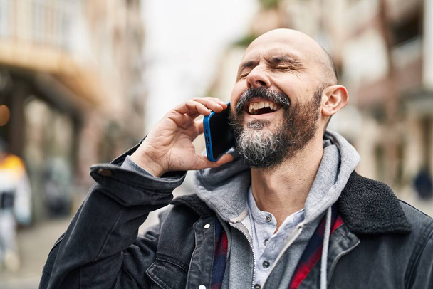 Νέοι φαλακρός άνθρωπος χαμογελά αυτοπεποίθηση μιλώντας στο smartphone στο δρόμο - Φωτογραφία, εικόνα