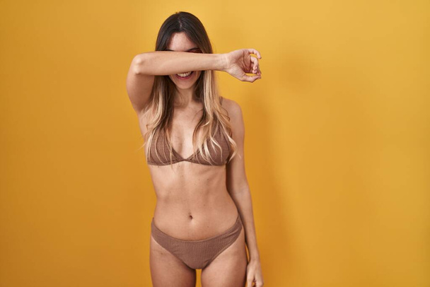 Young hispanic woman wearing bikini over yellow background covering eyes with arm smiling cheerful and funny. blind concept.  - Photo, Image