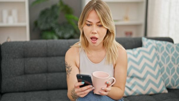 Nuori blondi nainen käyttää älypuhelin juo kahvia yllätys ilme kotona - Valokuva, kuva