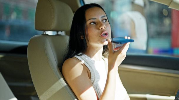 Young beautiful hispanic woman sending voice message by smartphone sitting on car at street - Photo, image