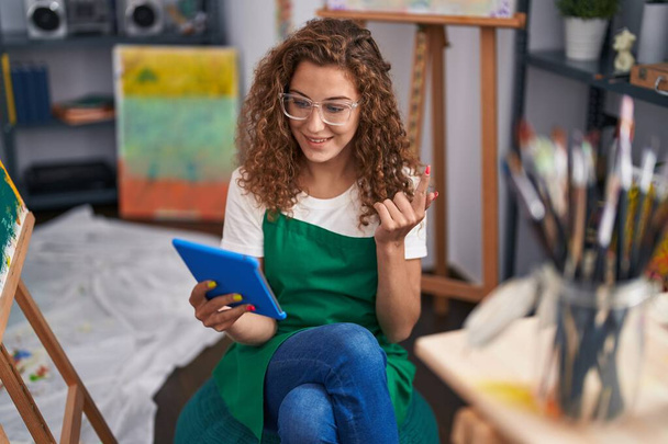 Young caucasian woman painter sitting at art studio doing video call with tablet smiling happy pointing with hand and finger to the side  - Photo, Image