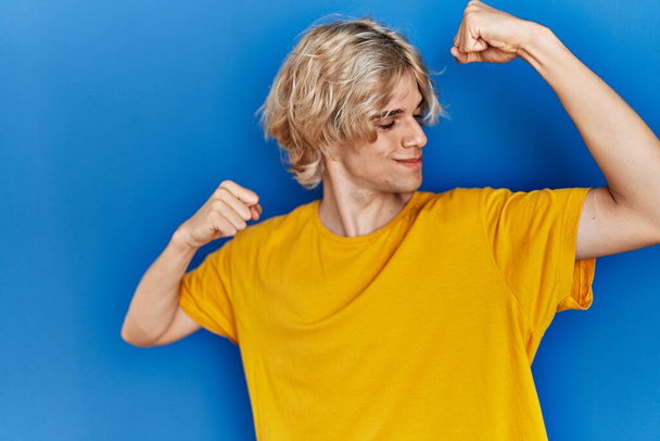 Jeune homme moderne debout sur fond bleu montrant les muscles des bras souriant fier. concept de fitness.  - Photo, image