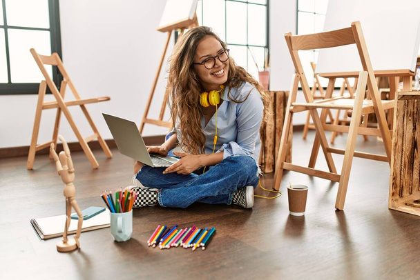 Young beautiful hispanic woman artist using laptop sitting on floor at art studio - Photo, Image