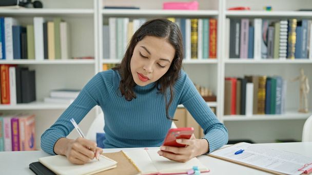 Jonge mooie Spaanse vrouw student met behulp van smartphone schrijven op notebook op de universiteit klaslokaal - Foto, afbeelding