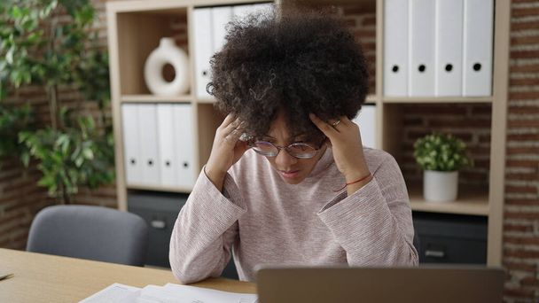 若いアフリカ系アメリカ人女性ビジネスワーカーがオフィスでストレスの多い表情でテーブルの上に座る - 写真・画像