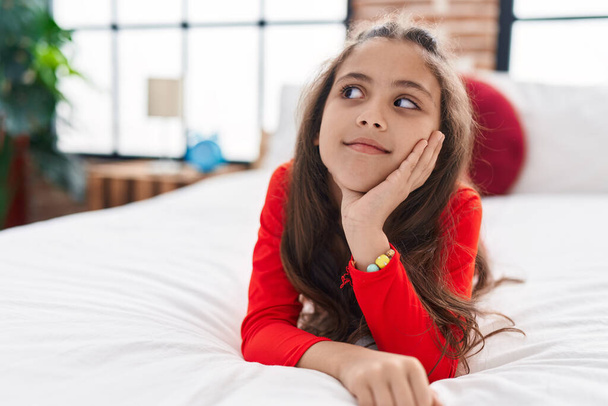 Adorable hispanic girl lying on bed with doubt expression at bedroom - Photo, Image