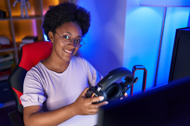 Африканская американка стример улыбается уверенно держа наушники в игровой комнате - Фото, изображение