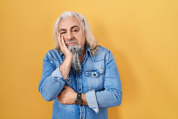 Mann mittleren Alters mit grauen Haaren steht vor gelbem Hintergrund und sieht müde und gelangweilt aus mit Depressionsproblemen mit verschränkten Armen.  - Foto, Bild