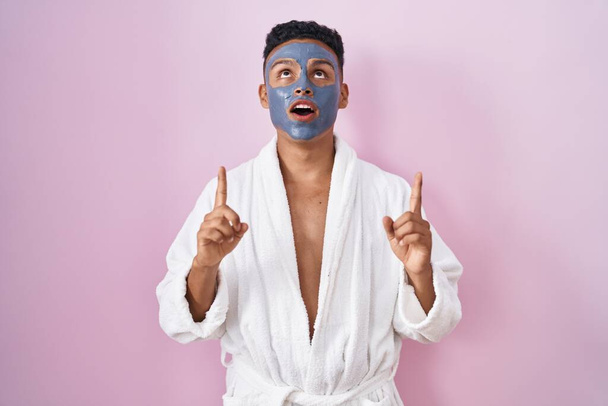 Jonge Latijns-Amerikaanse man draagt schoonheid gezichtsmasker en badjas verbaasd en verrast omhoog te kijken en te wijzen met vingers en opgeheven armen.  - Foto, afbeelding