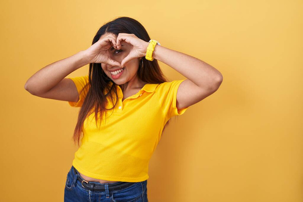 Young arab woman standing over yellow background doing heart shape with hand and fingers smiling looking through sign  - Fotó, kép