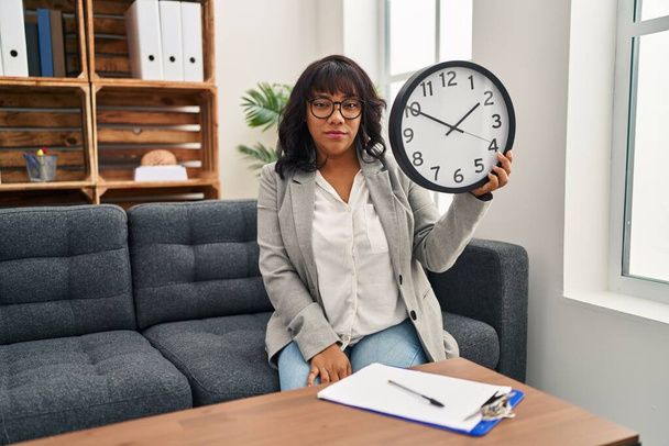 Hispanic woman working at therapy office holding clock thinking attitude and sober expression looking self confident  - Fotoğraf, Görsel