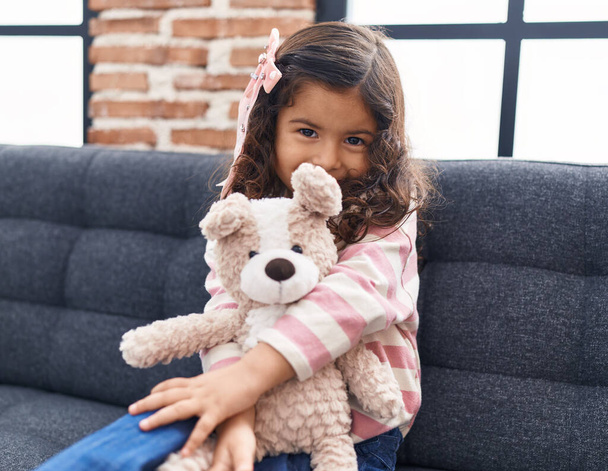 Schattige latino meisje knuffelen teddy beer zitten op de bank thuis - Foto, afbeelding