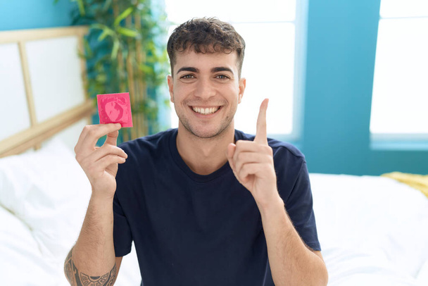 Joven hispano sentado en la cama sosteniendo condón sonriendo feliz señalando con la mano y el dedo hacia un lado  - Foto, Imagen