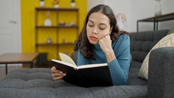 Jovem bela mulher hispânica lendo livro deitado no sofá em casa - Foto, Imagem
