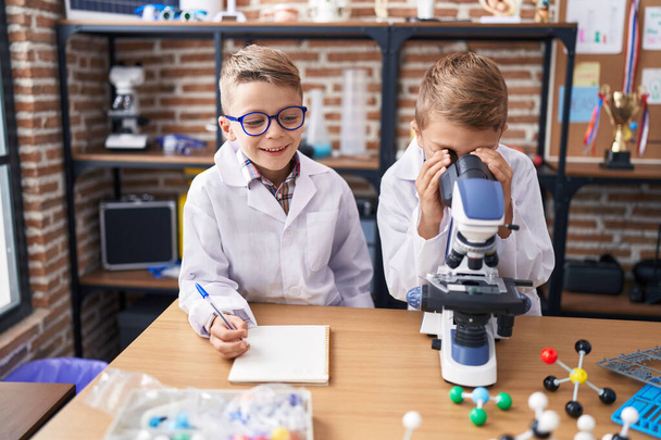 Uroczy chłopcy studenci korzystający z mikroskopu pisanie notatek w klasie laboratoryjnej - Zdjęcie, obraz