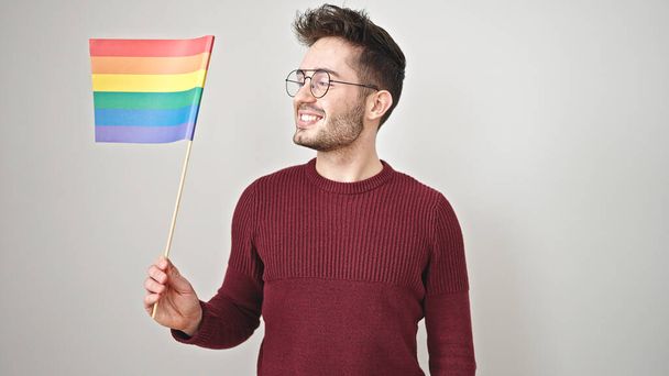 Giovane uomo ispanico sorridente fiducioso tenendo bandiera arcobaleno su sfondo bianco isolato - Foto, immagini