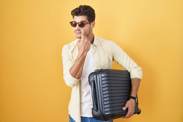 Młody Latynos trzymający walizkę na wakacjach, wskazujący oko na twój gest, podejrzany wyraz twarzy.  - Zdjęcie, obraz