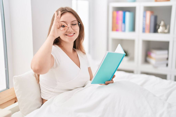 Young caucasian woman sitting on the bed at home reading a book smiling happy doing ok sign with hand on eye looking through fingers  - Fotoğraf, Görsel