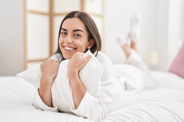 Young hispanic woman hugging pillow lying on bed at bedroom - Foto, Imagen